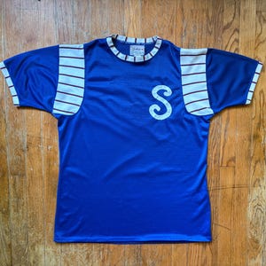 Może przedstawiać: Winylowa niebieska koszulka z siatki z białymi i bordowymi paskami na rękawach. Koszulka ma białą literę "S" z przodu oraz biało-czarny kołnierz w paski. Koszula leży płasko na drewnianej powierzchni.