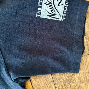 May include: Black short-sleeved t-shirt with a white Nautica logo on the sleeve. The logo says "The Power" and "Nautica" with a sailboat graphic. The text "THE FLATS-C" is also on the sleeve.