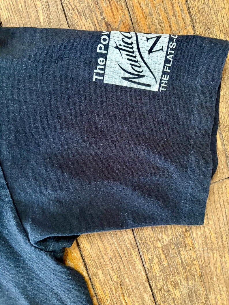May include: Black short-sleeved t-shirt with a white Nautica logo on the sleeve. The logo says "The Power" and "Nautica" with a sailboat graphic. The text "THE FLATS-C" is also on the sleeve.
