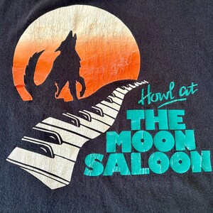 May include: Black t-shirt with a graphic of a wolf howling at a moon with a keyboard in the foreground. The text "Howl at The Moon Saloon" is printed in green.
