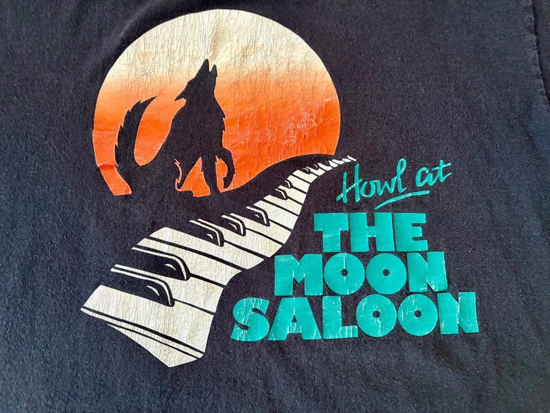 May include: Black t-shirt with a graphic of a wolf howling at a moon with a keyboard in the foreground. The text "Howl at The Moon Saloon" is printed in green.