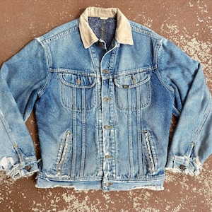 May include: A blue denim jacket featuring a tan corduroy collar, a button-up front, two chest pockets with button flaps, and two lower vertical pockets. The jacket displays visible fraying and distressed wear along the cuffs and hemline.
