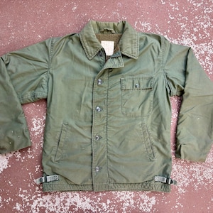 May include: An olive green long-sleeve jacket featuring a pointed collar, a button-front closure, and a single chest pocket with a button flap. The jacket includes two lower slash pockets and adjustable side straps with metal buckles at the hem.