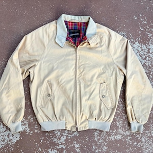 Peut inclure: Une veste zippée beige dotée d'un col clair, de manches longues avec poignets côtelés et d'un ourlet côtelé. La doublure intérieure présente un motif à carreaux rouge, vert et bleu. Deux poches avant avec fermetures à boutons sont visibles.