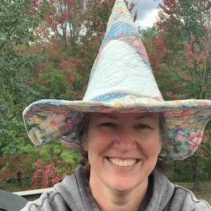 May include: A tall, pointed hat made of quilted fabric featuring a floral pattern in shades of blue, pink, and white. The hat has a wide, flexible brim and a conical crown, constructed from multiple fabric patches stitched together.