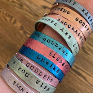 May include: A stack of seven colorful metal bracelets with different phrases engraved on them, including "Knit Happy", "Justing Goddess", "Procrastinate", "Wander Are Lost", "Yarn Goddess Blessed", "Unwind", and "Yarn Goddess Wish".