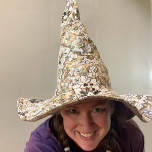 May include: A tall, pointed hat featuring a beige, brown, and green floral pattern. The hat has a wide, slightly curved brim and a structured, conical crown. The fabric displays a detailed, repeating botanical print with dark outlines.