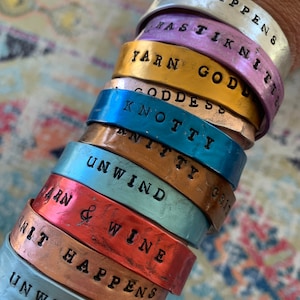 May include: A stack of eight colorful metal bracelets with different sayings stamped on them. The sayings include "Knit Happens", "Yarn God", "Knotty", "Unwind", "Yarn & Wine", and "It Happens".