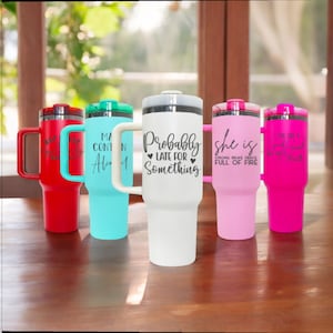 May include: Five stainless steel travel mugs with different designs and colors. The mugs have handles and lids. The designs include "Probably Late For Something", "She is Strong Brave Fierce Full of Fire", "I've Got A Good Heart But I'm Not Afraid To Use It", "Ma Contin Ale", and a red mug with a design that is not visible.