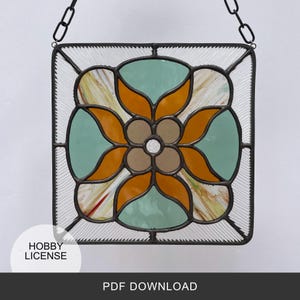 May include: A stained glass window panel with a geometric design featuring orange, green, and brown glass pieces. The panel is framed in black metal and has a chain for hanging.