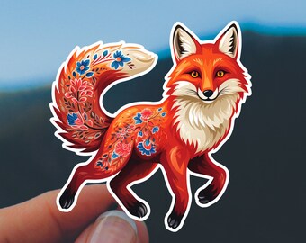 May include: A vibrant sticker featuring a stylized fox with a red and orange coat, adorned with floral patterns in blue, pink, and white. The fox has a white chest and a fluffy tail. The sticker is held against a backdrop of a mountain range and a blue sky.