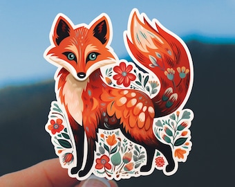 May include: A colorful sticker featuring a stylized fox with orange and white fur, blue eyes, and a bushy tail. The fox is surrounded by flowers and foliage in shades of red, orange, and green. The sticker is held against a backdrop of a blue sky and mountains.