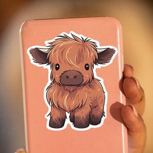 May include: A sticker featuring a cartoon illustration of a Highland cow calf. The calf has brown fur, dark brown hooves, and large, expressive eyes. The sticker is affixed to a pink phone case.