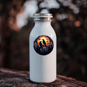 Puede incluir: Una botella de agua azul claro con tapa plateada. La botella presenta una pegatina circular con la silueta de dos p&aacute;jaros posados en una rama, sobre un atardecer con &aacute;rboles.