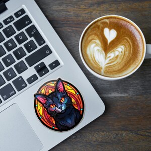 May include: A circular sticker with a black cat design, orange eyes, and pink ears, set against a stained-glass style background of orange, yellow, and red. The sticker is on a laptop next to a coffee cup with latte art.