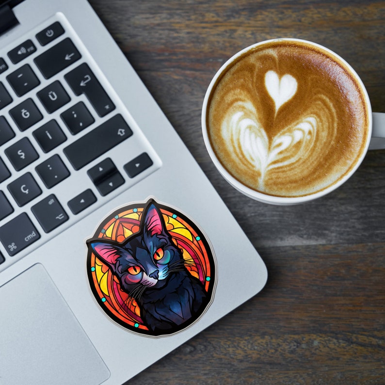 May include: A circular sticker with a black cat design, orange eyes, and pink ears, set against a stained-glass style background of orange, yellow, and red. The sticker is on a laptop next to a coffee cup with latte art.