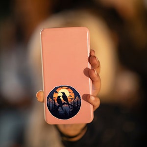 Puede incluir: Una pegatina redonda con una escena de puesta de sol con dos p&aacute;jaros negros posados en una rama. El fondo incluye un sol naranja, &aacute;rboles silueteados y un cielo azul oscuro. La pegatina est&aacute; adherida a un smartphone rosa.