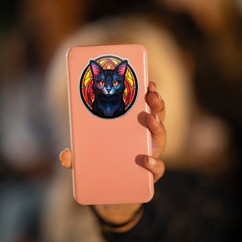May include: A sticker depicting a black cat with orange and blue eyes, in a stained-glass design. The sticker is on a pink phone, held in a hand. The cat design has a circular border.