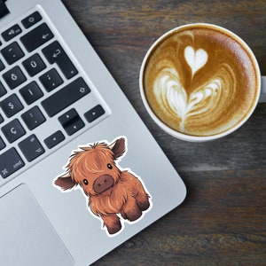 May include: A sticker of a cartoon Highland cow with brown fur, large ears, and a cute expression. The sticker is on a laptop next to a cup of coffee with latte art.