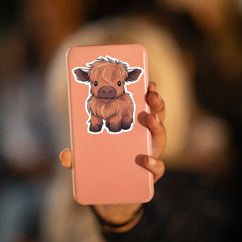 May include: A sticker featuring a cartoon illustration of a Highland cow calf. The calf has brown fur, dark brown hooves, and large, expressive eyes. The sticker is affixed to a pink phone case.