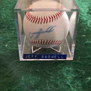 May include: A white baseball with red stitching, featuring a blue ink autograph, enclosed in a clear square acrylic display case. A blue nameplate at the base of the case displays the white text JEFF BAGWELL in all capital letters.