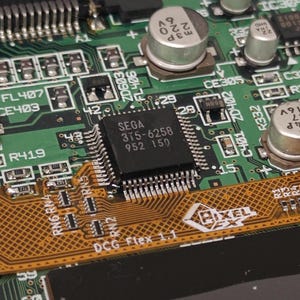 May include: Close-up of a green circuit board with a black chip labeled "SEGA 375-6258 952 150". The chip is surrounded by various electronic components, including capacitors and resistors.