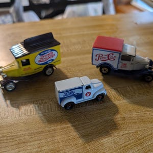 May include: Three vintage Pepsi-Cola delivery trucks in yellow, white, and blue. The trucks have detailed features, including the Pepsi-Cola logo and text. The trucks are on a wooden surface.