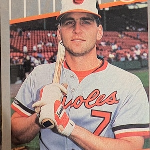 May include: A baseball card featuring Bill Ripken, a second baseman for the Baltimore Orioles. The card shows Ripken in his team uniform, holding a baseball bat. The card has the Fleer logo.