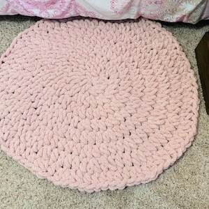 May include: A round, blush pink, chunky knit rug. The rug is made of thick, soft yarn and has a textured surface. The rug is placed on a neutral-colored carpet. The rug is approximately 36 inches in diameter.