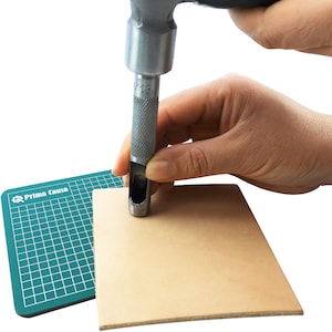 May include: A hand using a metal leather punch tool to make a hole in a piece of tan leather. The leather is on top of a green cutting mat with the text "Prima Causa" printed on it.