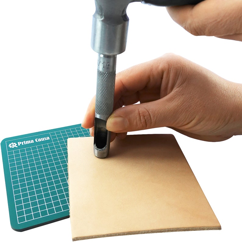 May include: A hand using a metal leather punch tool to make a hole in a piece of tan leather. The leather is on top of a green cutting mat with the text "Prima Causa" printed on it.