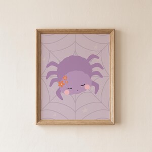 May include: A framed wall art print featuring a lavender spider with closed eyes, pink cheeks, and small orange flowers on its head. The spider is centered on a light purple background with a subtle spiderweb pattern and faint white stars.