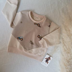 Könnte beinhalten: Ein beiger Langarmsweatshirt mit einem braunen und weißen Traktor-Druck. Der Pullover hat kontrastierenden beigefarbenen Stoff an den Ärmeln und eine diagonale Naht auf der Vorderseite.