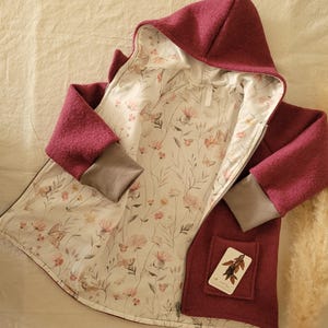 Könnte beinhalten: Burgunderfarbene Kapuzenjacke mit floralem und Schmetterlings-Print-Futter. Die Jacke hat einen Reißverschluss, graue Manschetten und eine kleine Tasche mit einem Etikett. Das Futter zeigt ein zartes Muster aus rosa Blumen und Schmetterlingen.
