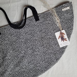 Könnte beinhalten: Eine schwarz-weiß Tragetasche mit Leopardenmuster. Die Tasche hat schwarze Griffe und ein Etikett mit einer botanischen Illustration. Die Tasche besteht aus einem weichen Material und eignet sich zum Tragen verschiedener Gegenstände.