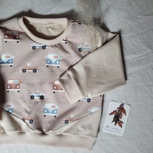 Könnte beinhalten: Beiges Langarm-Sweatshirt mit einem Muster aus braunen und blauen Cartoon-Autos. Die Ärmel und der Kragen sind einfarbig beige. Ein kleines Etikett mit Text und einem Blattdesign ist sichtbar.
