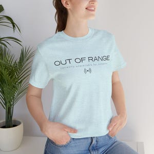Pode incluir: Uma t-shirt de manga curta em azul claro mesclado com o texto preto OUT OF RANGE, a frase currently unavailable for nonsense e um ícone de sinal. A peça possui gola redonda e um corte padrão, oferecendo um visual casual e descontraído.