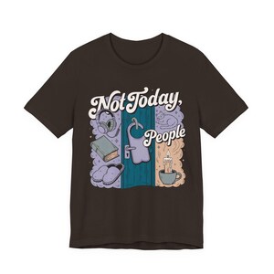 May include: A dark brown t-shirt featuring a graphic print with the text "Not Today, People" above a blue door. The illustration includes a pair of headphones, a book, slippers, a sleeping cat, and a lit candle in a mug, all in soft pastel tones.