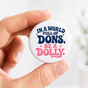 May include: A round white pin-back button featuring dark blue text that reads IN A WORLD FULL OF DONS, and pink text that reads BE A DOLLY. The design includes small star and flower accents, creating a clear and legible graphic on the circular surface.