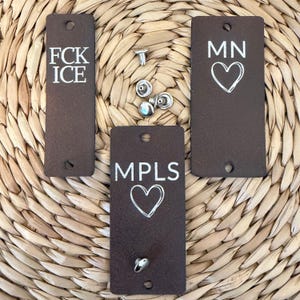 May include: Three rectangular brown leather tags with white text and heart designs. One tag reads "FCK ICE", another "MN" with a heart, and the third "MPLS" with a heart. Several silver rivets and fasteners are scattered around the tags.