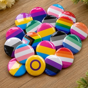 May include: A collection of colorful round buttons, each featuring a different design representing various LGBTQ+ identities. The buttons display stripes, gradients, and symbols in a variety of colors, including pink, blue, yellow, and green. The buttons are arranged on a wooden surface.