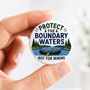 May include: A round white pin-back button featuring a graphic of a dark green canoe on blue water with pine trees. The text reads PROTECT THE BOUNDARY WATERS and NOT FOR MINING in dark green and navy blue lettering with small pine tree icons.