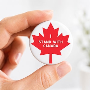 May include: A circular white pin-back button featuring a large red maple leaf graphic. The text "I STAND WITH CANADA" is printed in white capital letters across the center of the red maple leaf design on the front of the button.