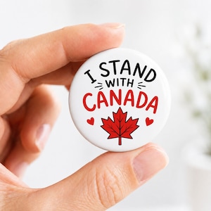 May include: A round white pin-back button featuring the black text I STAND WITH and the red text CANADA. Below the text is a red maple leaf graphic flanked by two small red hearts. The design is centered on the circular surface of the button.