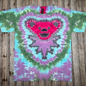 May include: A tie-dye t-shirt featuring a vibrant bear face design. The shirt showcases a spiral pattern in shades of pink, blue, green, and purple. The bear's face is a bright pink with black accents, creating a psychedelic and colorful aesthetic.