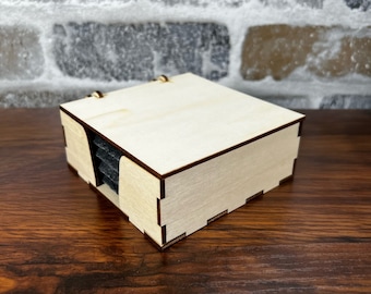 Puede incluir: Una caja de madera con una pila de posavasos negros en su interior. La caja está hecha de madera clara y tiene un diseño sencillo.