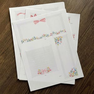 May include: A stack of white paper sheets with floral and bow illustrations. The designs include pink bows, floral borders, and corner accents in pastel colors. The papers are arranged on a dark wood surface.