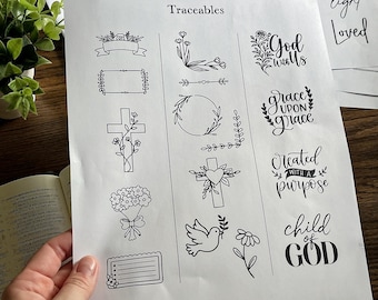 May include: A white sheet of paper titled Traceables featuring black line art illustrations including floral borders, a cross, a heart, a dove, and religious phrases such as God with us, grace upon grace, created with a purpose, and child of God.