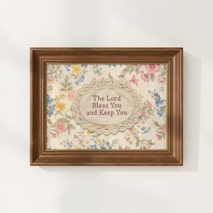 May include: A framed floral print with the text "The Lord Bless You and Keep You." The print features a cream-colored background with a delicate floral pattern in pink, blue, and yellow. The frame is a warm brown color.