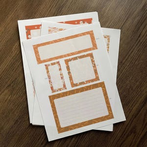 May include: A stack of white paper sheets with orange and floral decorative borders. The sheets feature various rectangular and square label designs, some with lined spaces for writing. The background is a dark wood surface.